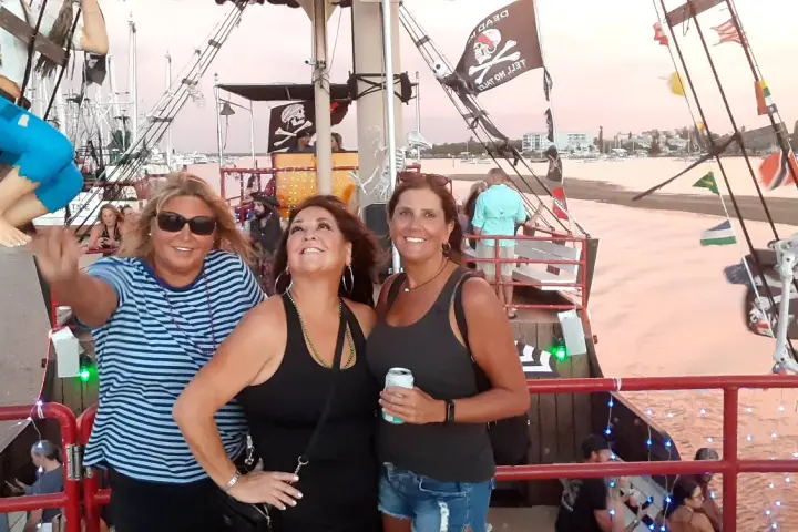 Three women pose on a pirate-themed boat at sunset with flags and decorations.