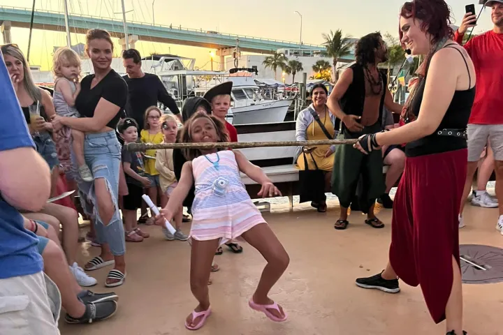 Child playing limbo outdoors, bending under a bar held by an adult, with onlookers in the background.