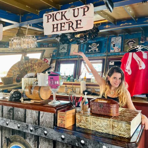 Woman at pirate-themed bar counter with 'Pick Up Here' sign overhead.