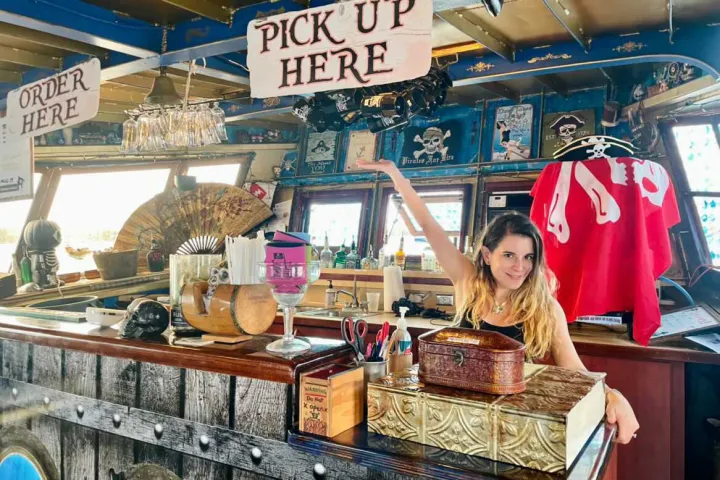Woman at pirate-themed bar counter with 'Pick Up Here' sign overhead.
