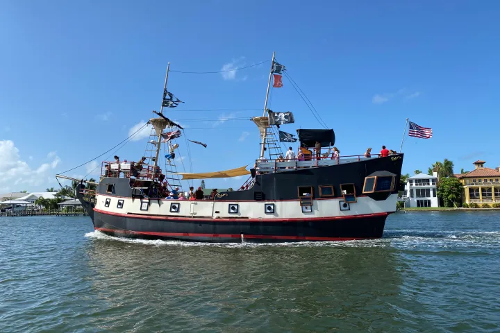 A pirate-themed boat with flags sailing on a sunny day near the coast.