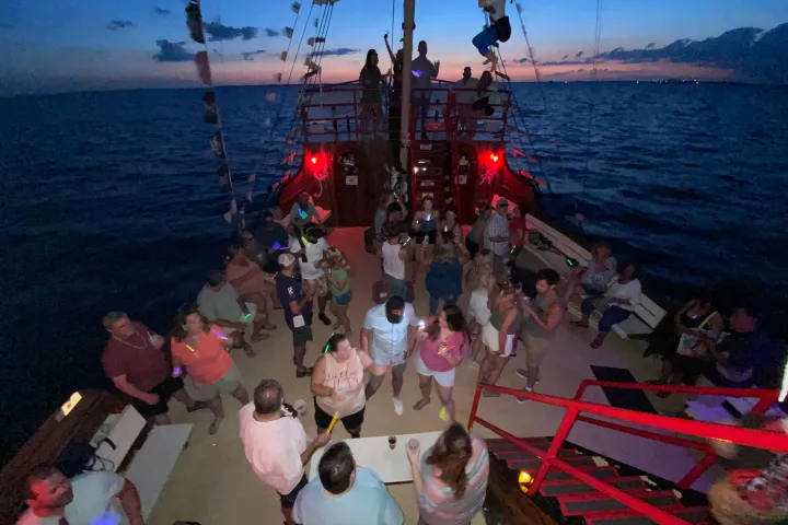 People partying on a ship deck at sunset, with red lights and open ocean in the background.