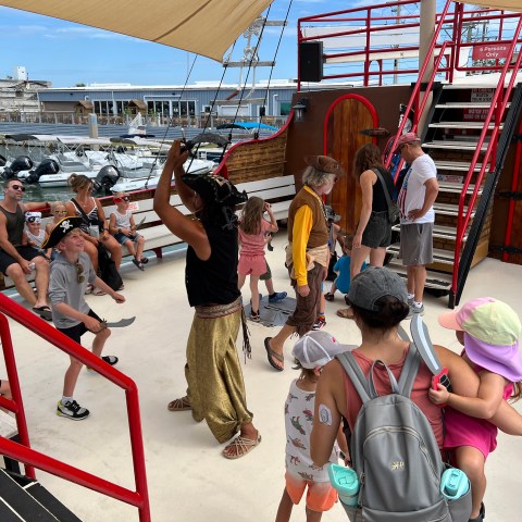 People in pirate costumes on a boat with children playing, dock visible in background.