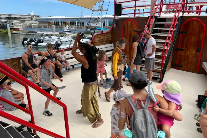 People in pirate costumes on a boat with children playing, dock visible in background.