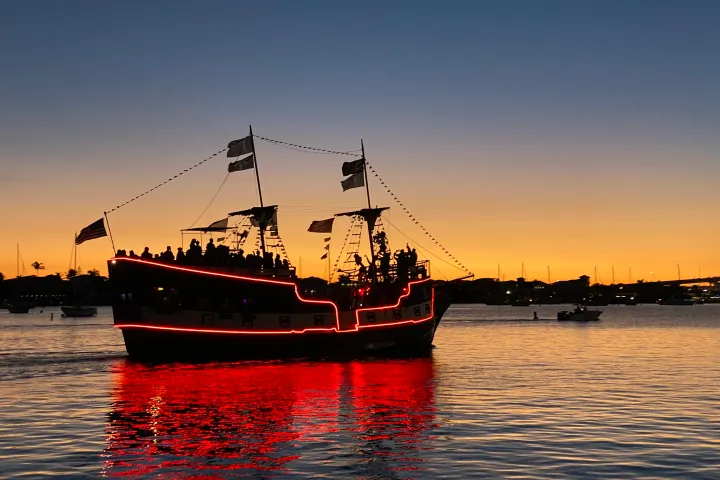 Pirate ship with red lights on water at sunset, silhouetted against orange sky.