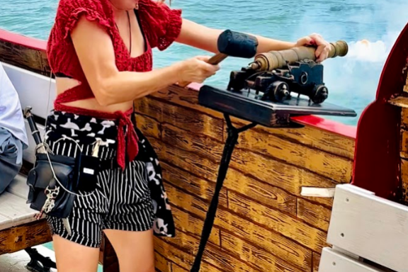 Person wearing ear protection using a small cannon on a wooden ship by the sea.