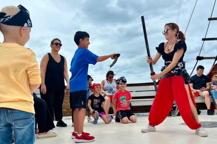 Children and a woman play with pirate costumes, smiling on a deck under a cloudy sky.