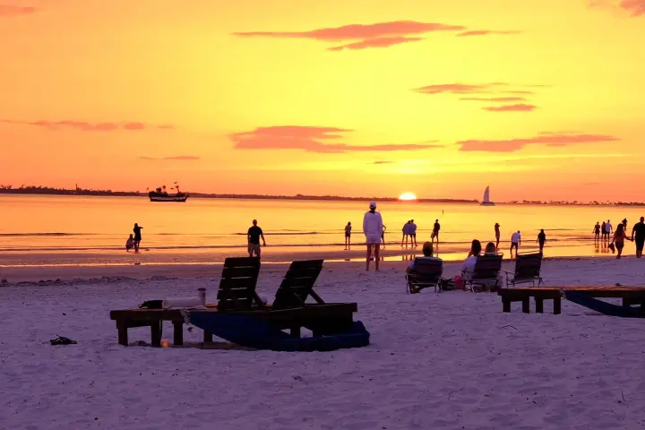 Beach scene at sunset with people, sunbeds, and boats on the horizon.