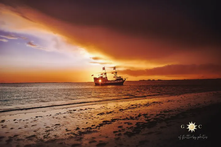 A ship sails at sunset, silhouetted against a vibrant sky on a tranquil beach.