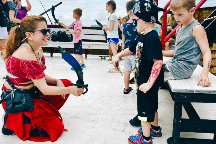 Woman in pirate costume interacting with children holding toy swords on a deck.