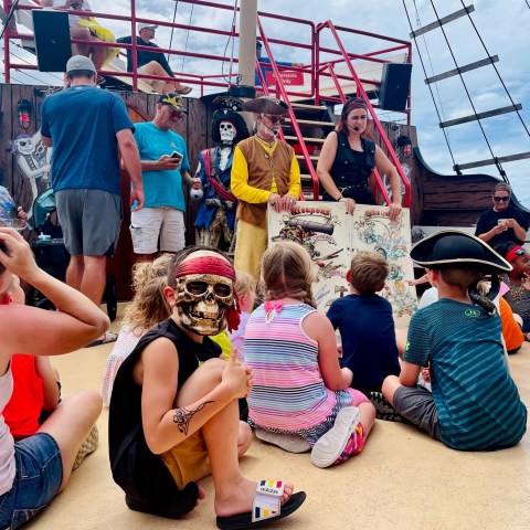 Children sit on a ship deck with pirates showing a treasure map.
