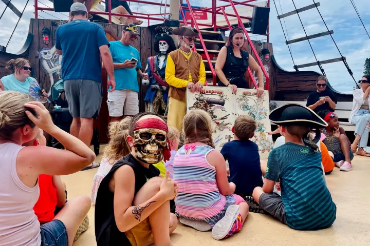 Children sit on a ship deck with pirates showing a treasure map.