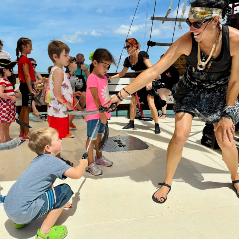 Children and adults in pirate costumes engage in a fun sword fight on a ship's deck.