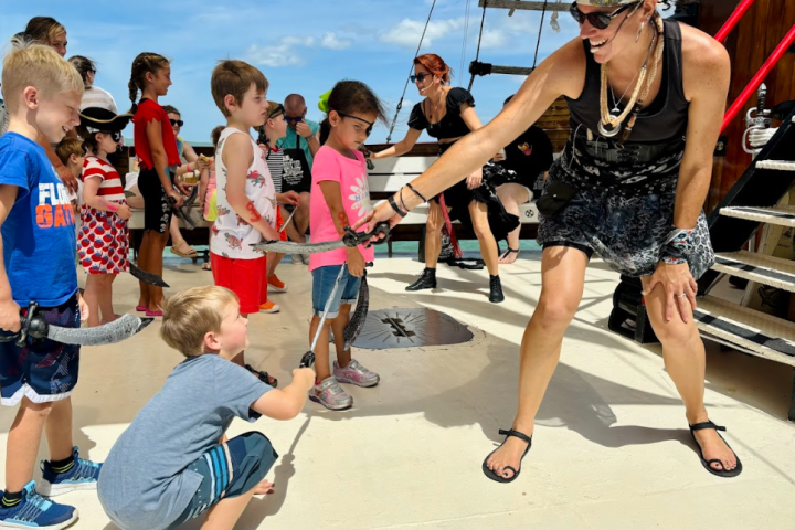 Children and adults in pirate costumes engage in a fun sword fight on a ship's deck.
