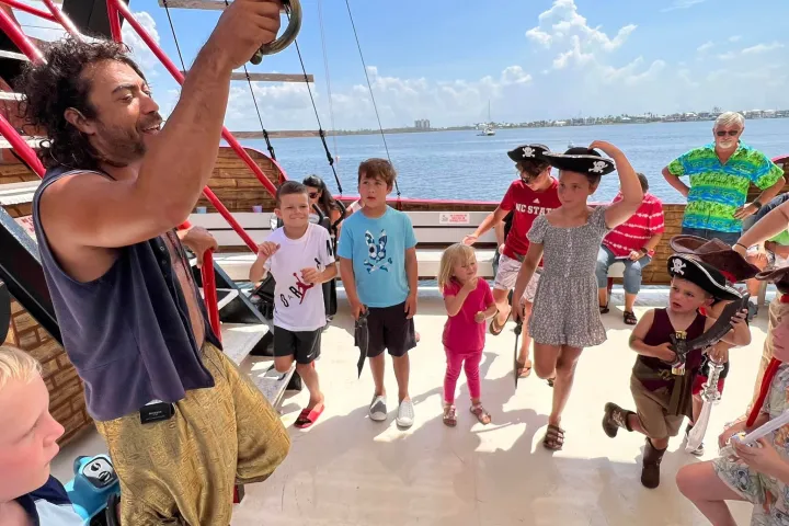 Group of kids on a boat with an adult dressed as a pirate holding a sword, sunny day, ocean in background.