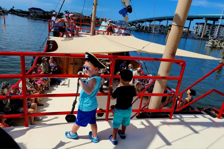 Two children on a boat deck with red rails, one wearing a pirate hat, with a sunny waterfront and bridge in view.