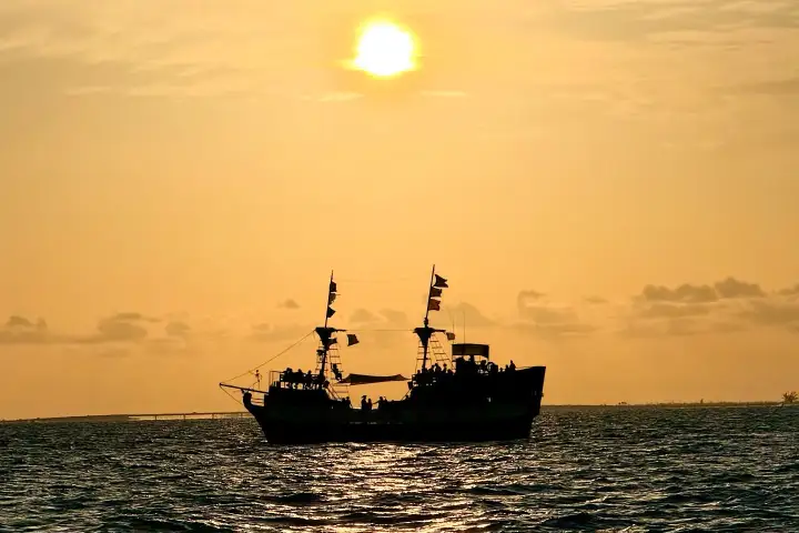 Silhouetted ship sailing at sunset with orange sky and calm sea.