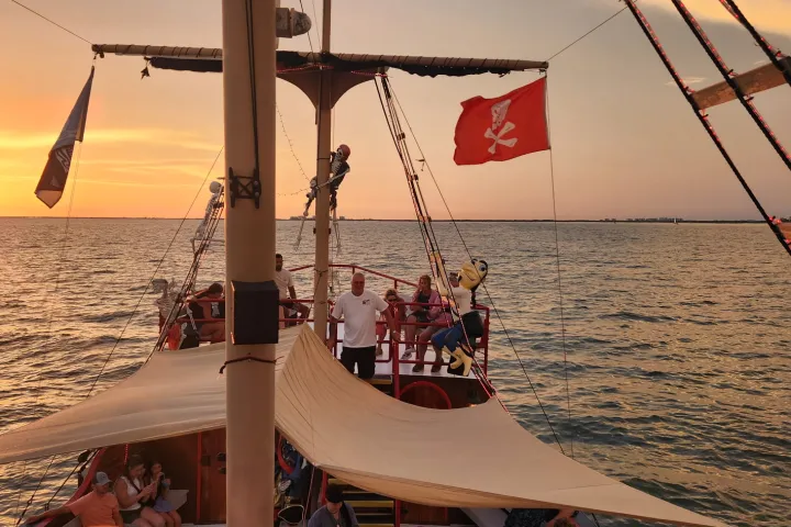 A pirate ship sails at sunset with people on deck under orange skies.