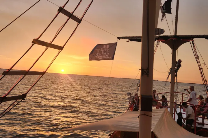 Pirate-themed ship at sunset with passengers and flags, sailing on a calm sea.