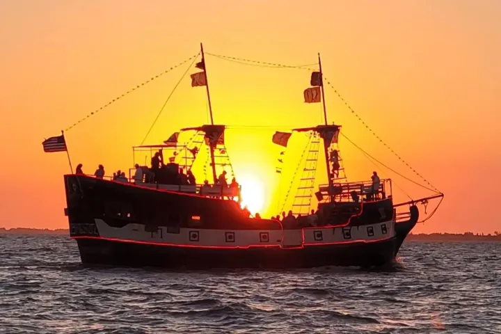 A pirate ship silhouette on the ocean at sunset with flags and people on board.