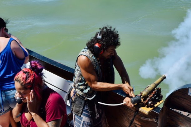 Person firing a small cannon on a boat, with others covering ears due to noise.