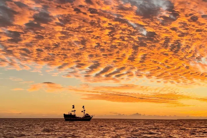Ship on ocean at sunset with vibrant orange clouds filling the sky.