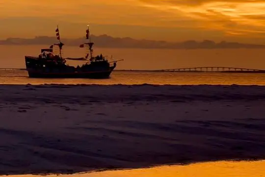 Silhouette of a ship at sunset with orange clouds over calm water reflection.