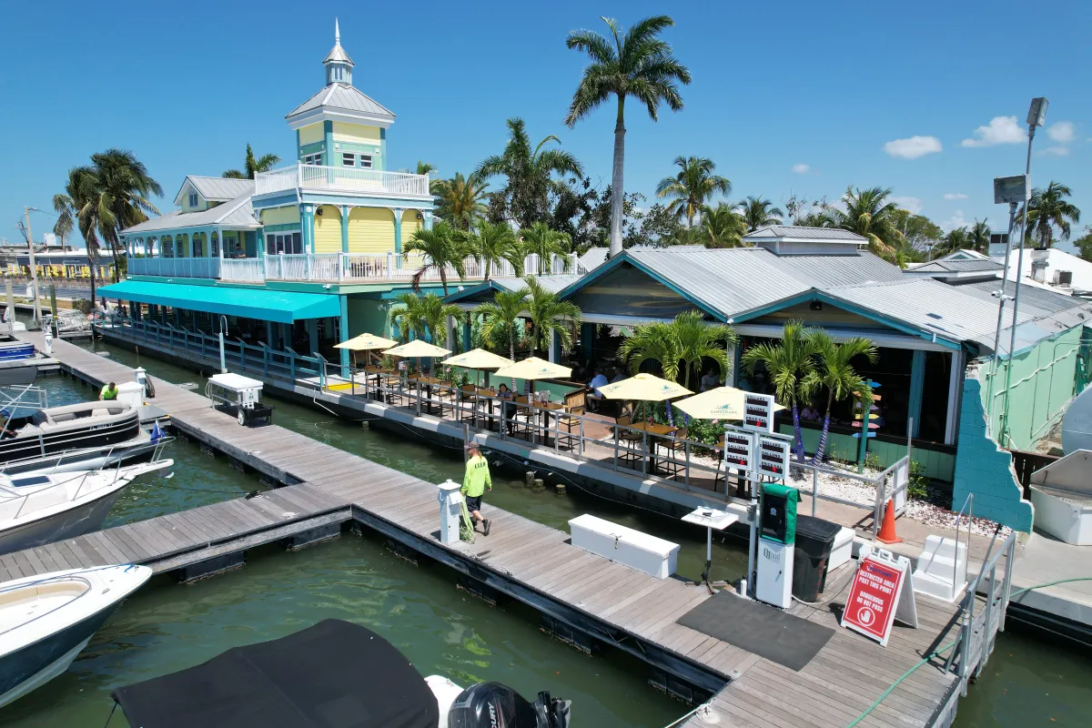 Waterfront restaurant with patio, umbrellas, palm trees, and docked boats.