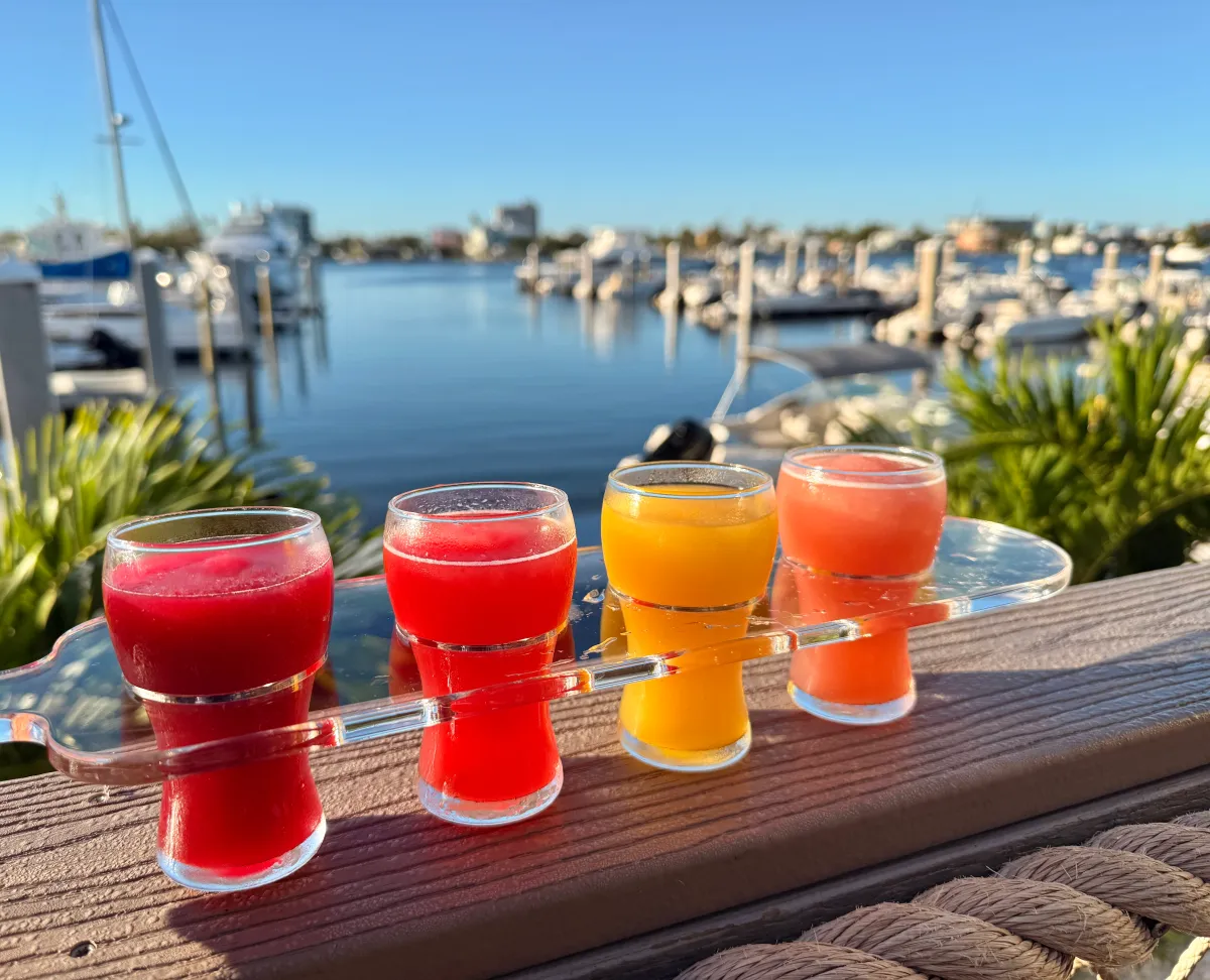Four colorful drinks in glasses overlooking a marina with boats and blue sky.
