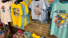 Display of colorful T-shirts and dresses with tropical and pirate-themed prints hanging and folded on shelves.