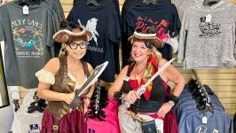 Two women in pirate costumes holding swords in front of a t-shirt display.