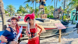 Two women in pirate costumes pose near a pirate statue and boat under palm trees.