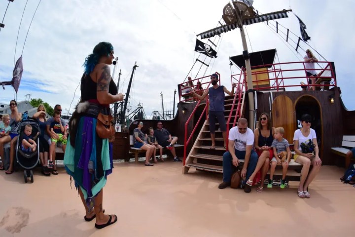 Tourists on a wooden ship, listening to a person dressed as a pirate, partially cloudy sky.