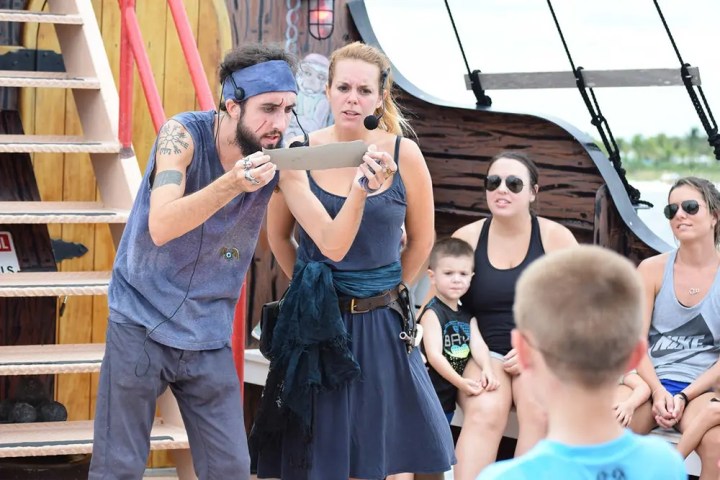 Man in pirate costume enthusiastically reads map to crowd on boat deck.
